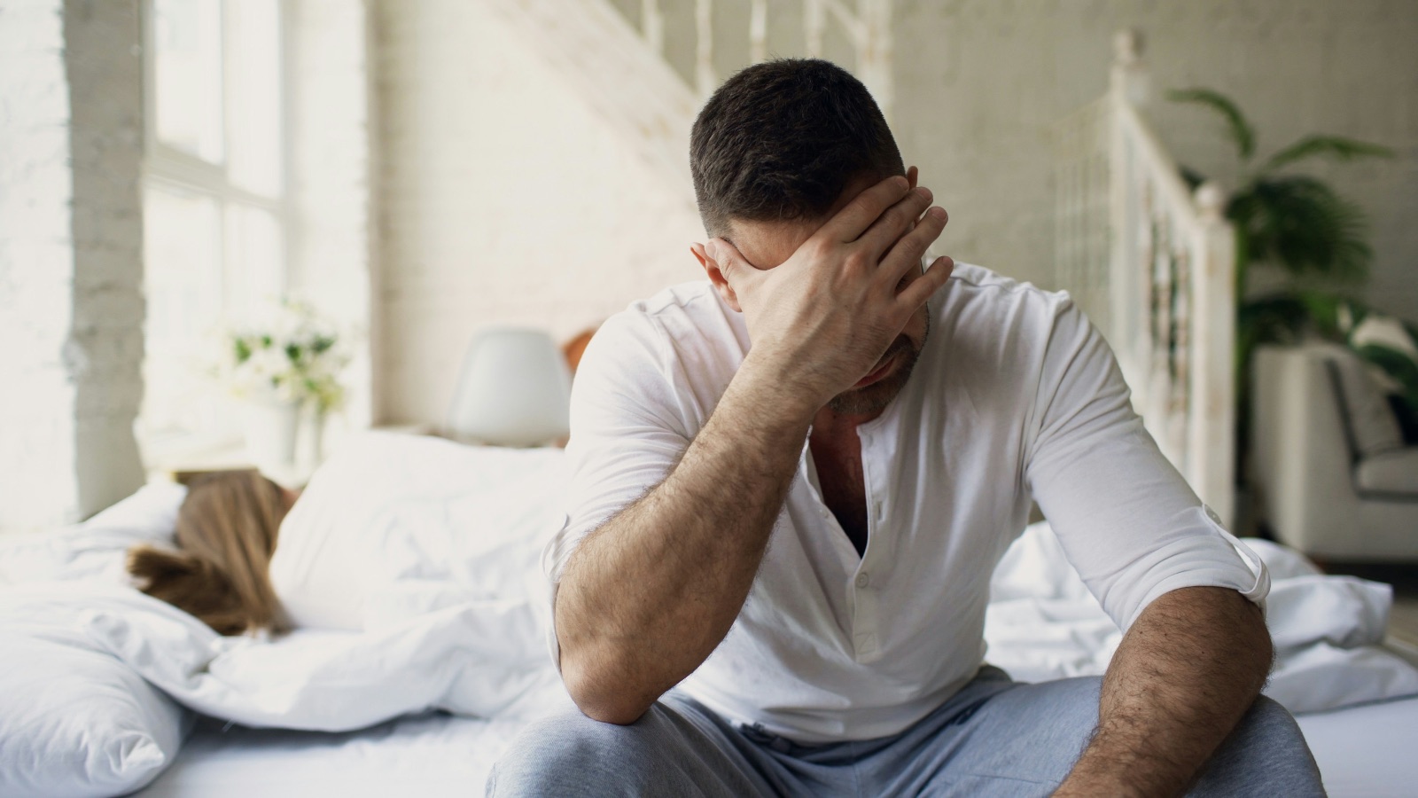 Man quietly sitting on the edge of the bed looking stressed and closed off emotionally.